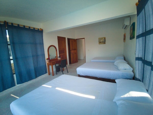 Recámara amplia con dos camas matrimoniales, cortinas azules, tocador de madera y aire acondicionado
