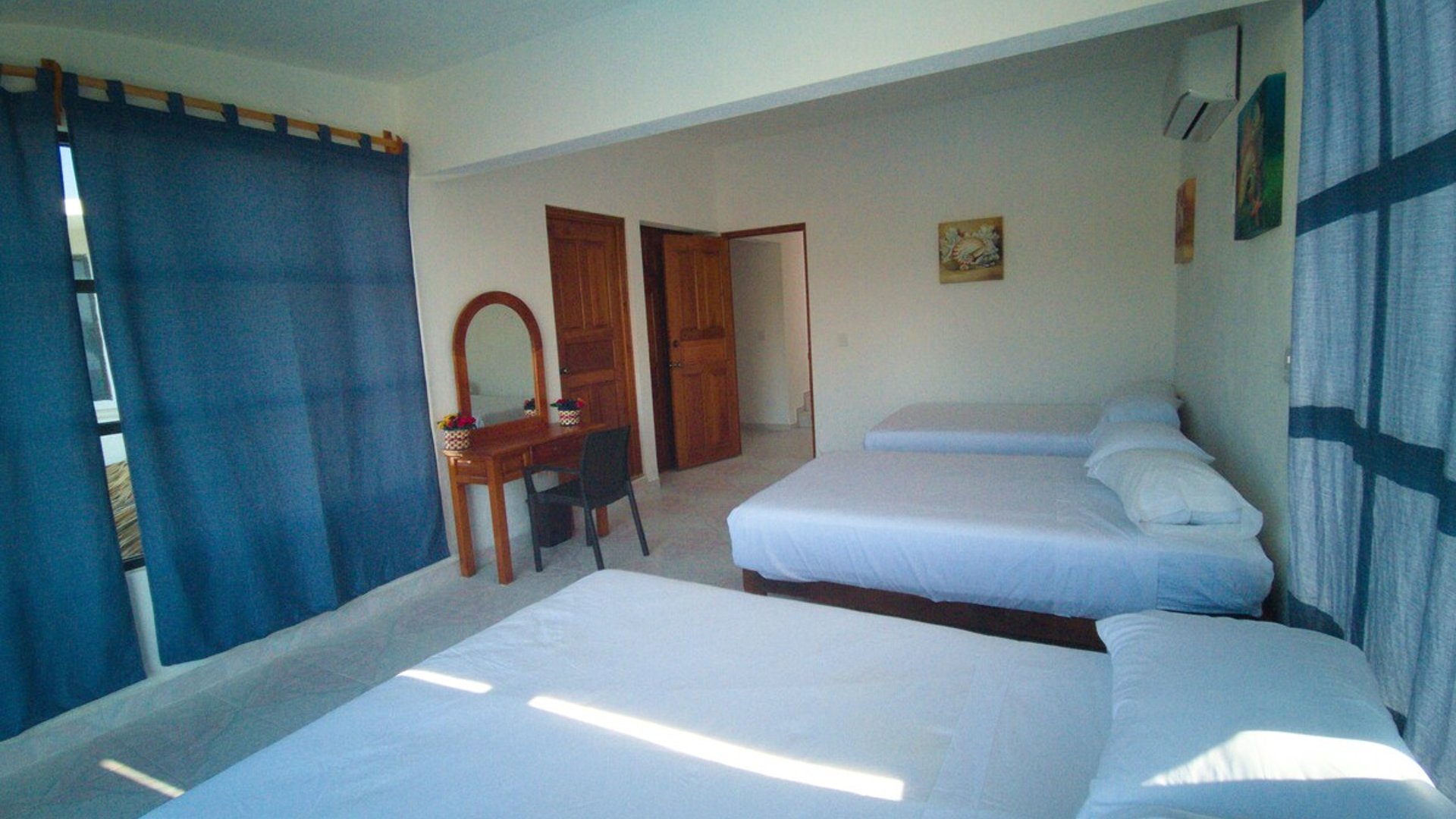 Recámara amplia con dos camas matrimoniales, cortinas azules, tocador de madera y aire acondicionado