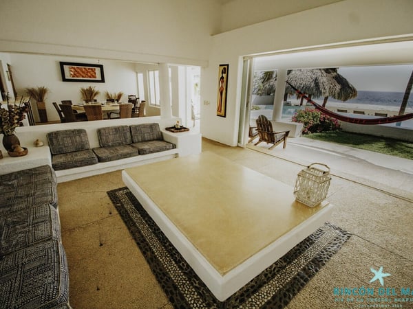 Sala abierta al mar con sofás grises, mesa de centro y vista a palapa, hamaca y océano al fondo