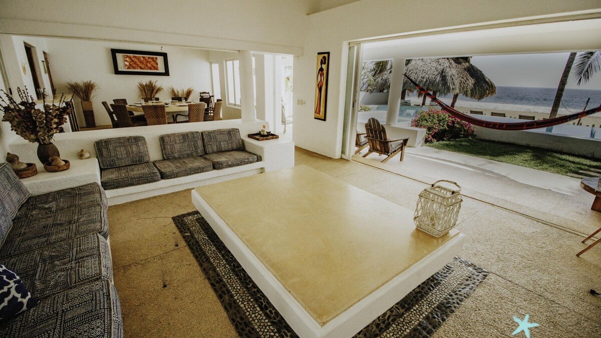 Sala abierta al mar con sofás grises, mesa de centro y vista a palapa, hamaca y océano al fondo
