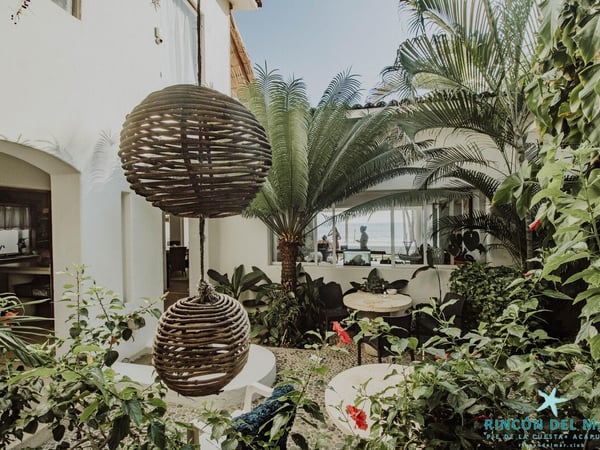 Jardín interior con esferas de mimbre colgantes, palmera cica y vegetación tropical; al fondo se asoma el mar