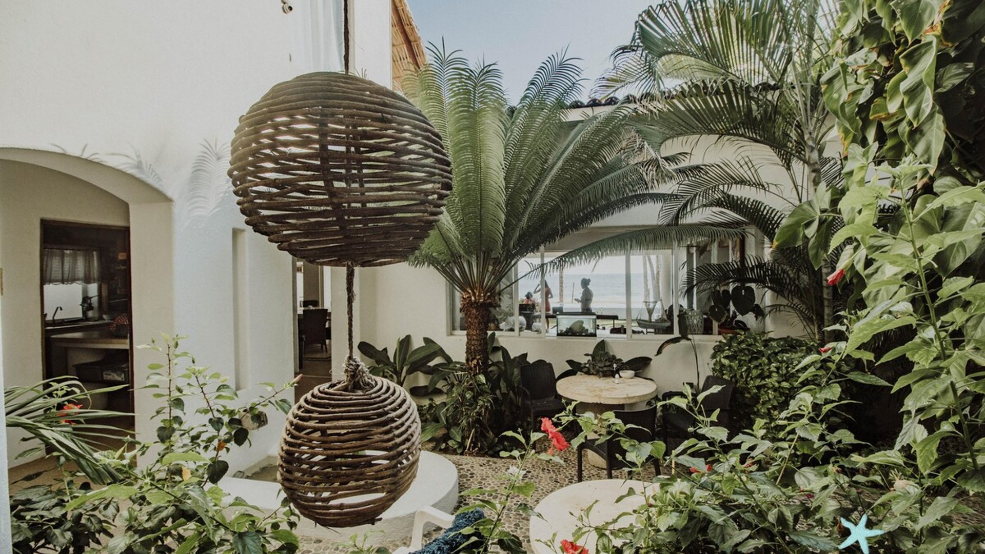 Jardín interior con esferas de mimbre colgantes, palmera cica y vegetación tropical; al fondo se asoma el mar