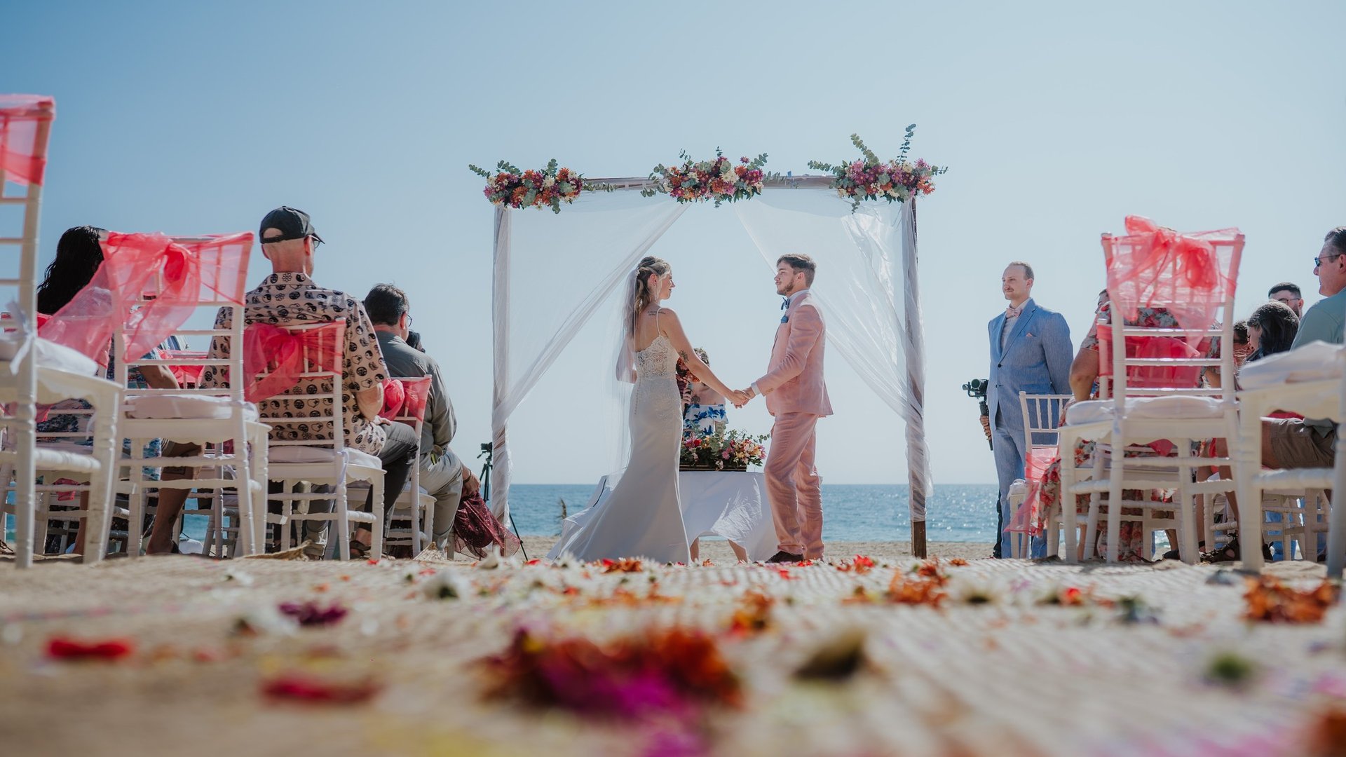 Ceremonia de boda en playa con arco floral y sillas decoradas frente al mar bajo cielo despejado