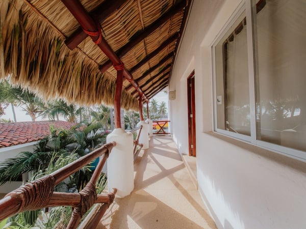 Corredor exterior con techo de palapa, barandal de madera y vistas al jardín tropical y al mar al fondo