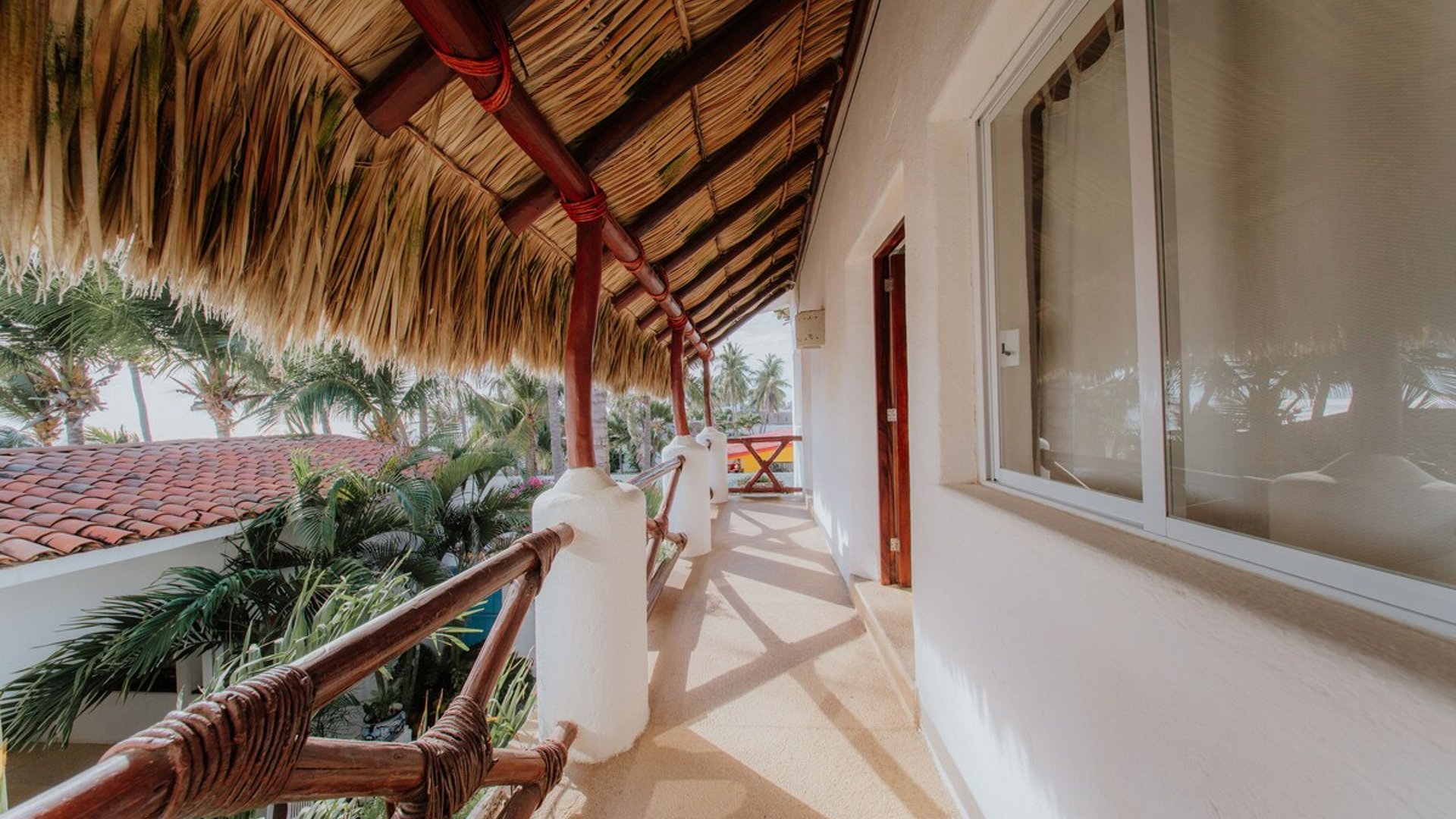 Corredor exterior con techo de palapa, barandal de madera y vistas al jardín tropical y al mar al fondo
