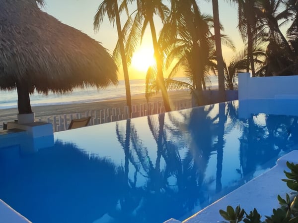 Alberca infinity frente al mar al atardecer, con palmeras y palapa enmarcando el sol sobre el Pacífico