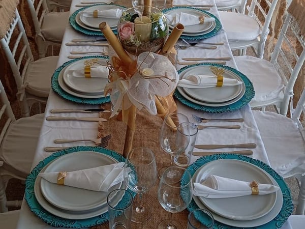 Mesa banquete con mantel blanco, platos turquesa y centros de bambú y flores para evento especial en terraza