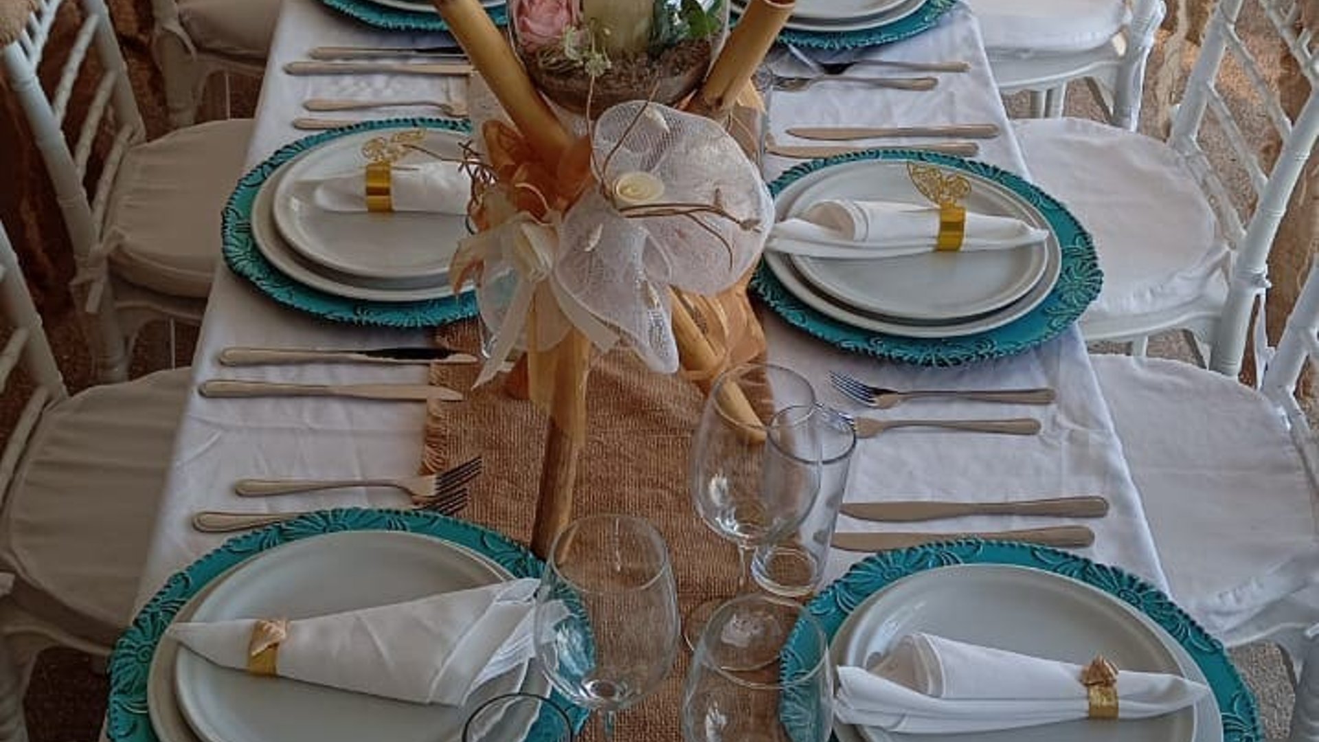 Mesa banquete con mantel blanco, platos turquesa y centros de bambú y flores para evento especial en terraza