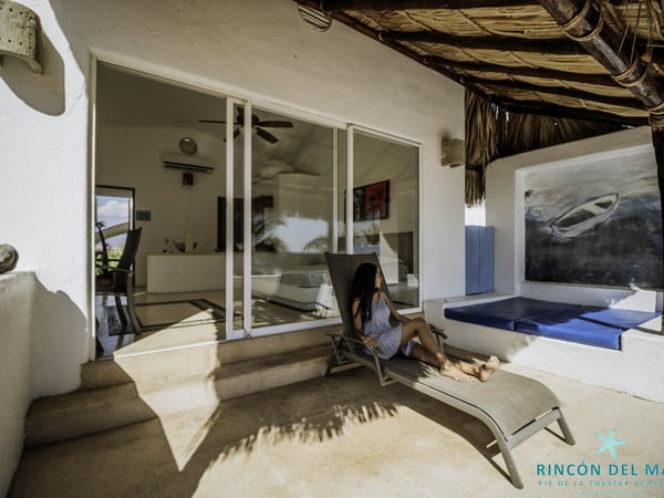 Terraza con camastro bajo palapa y vista al interior de recámara con ventanales deslizantes