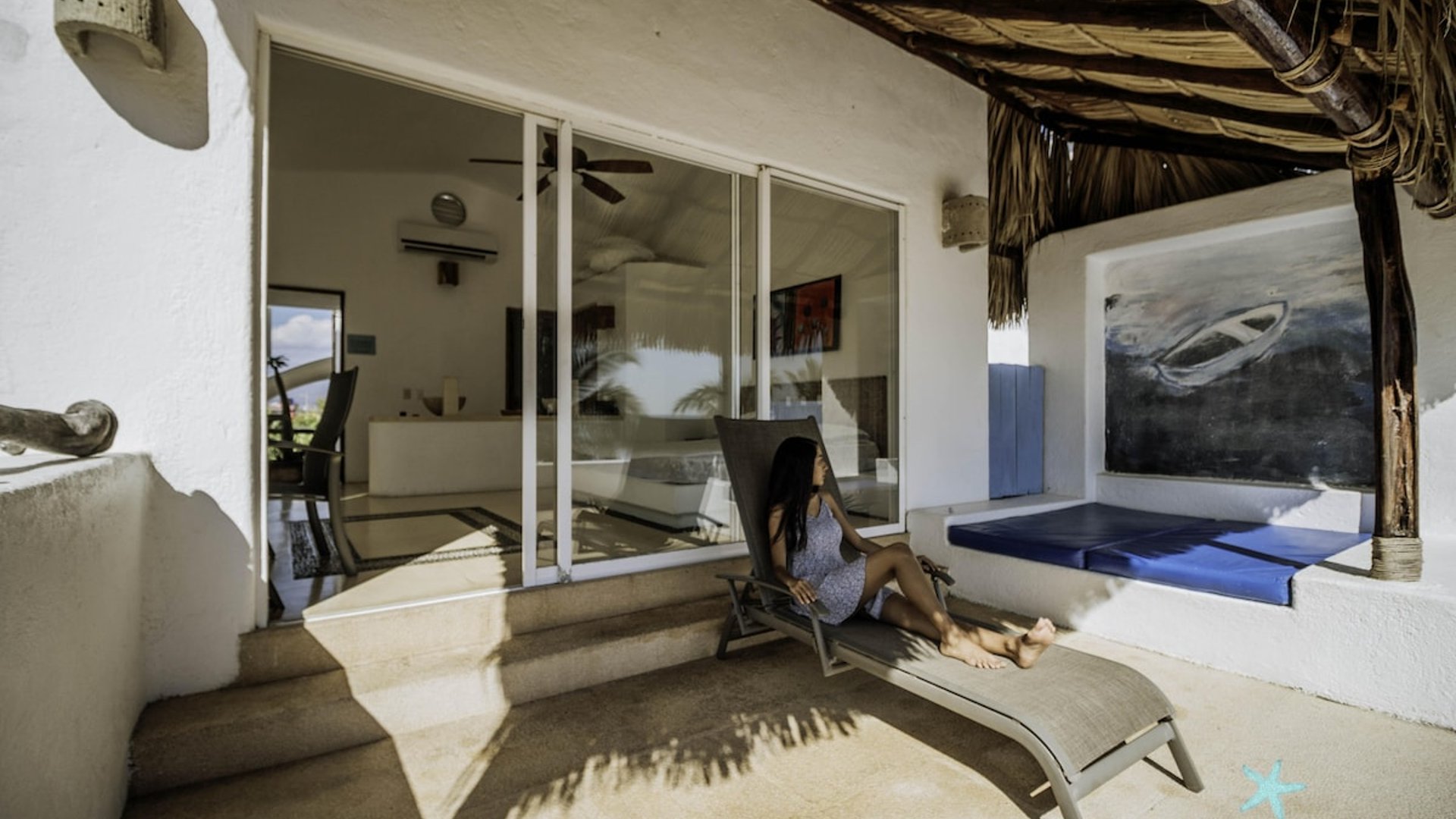 Terraza con camastro bajo palapa y vista al interior de recámara con ventanales deslizantes
