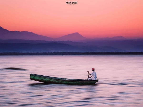 Pescador en canoa sobre laguna tranquila al atardecer con cielo rosa y silueta de montañas al fondo