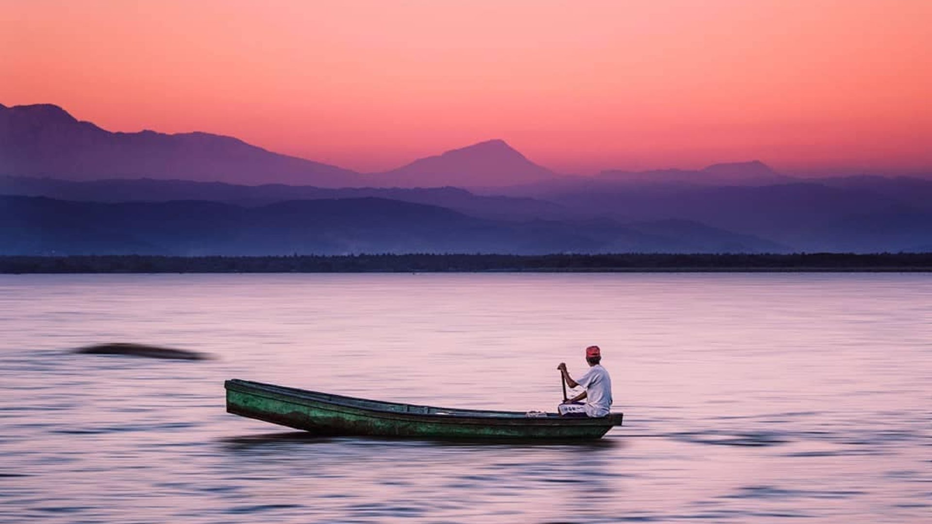 Pescador en canoa sobre laguna tranquila al atardecer con cielo rosa y silueta de montañas al fondo