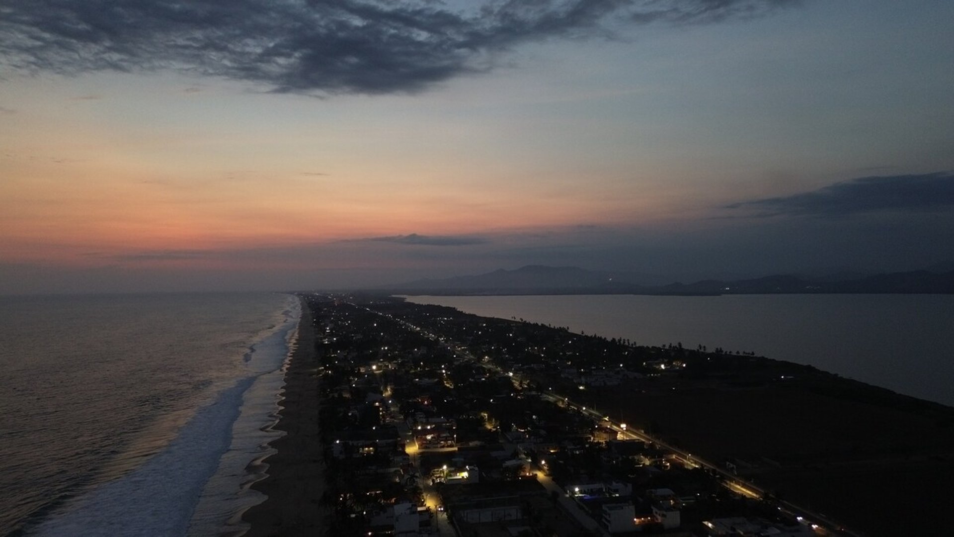 Vista aérea de Pie de la Cuesta al atardecer: franja de tierra entre el Pacífico y la laguna, luces encendidas bajo cielo rosado
