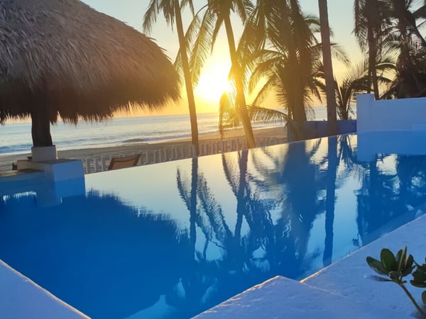 Alberca de borde infinito con vista al mar y palmeras al atardecer desde palapa frente a la playa