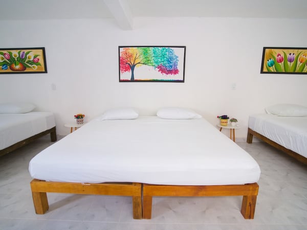 Recámara amplia con cama doble y dos camas individuales, paredes blancas decoradas con cuadros coloridos de flores y árbol arcoíris