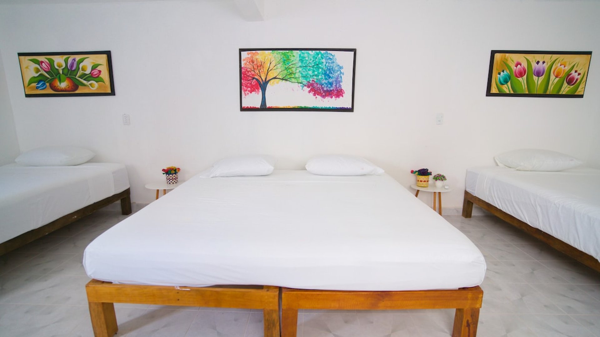 Recámara amplia con cama doble y dos camas individuales, paredes blancas decoradas con cuadros coloridos de flores y árbol arcoíris