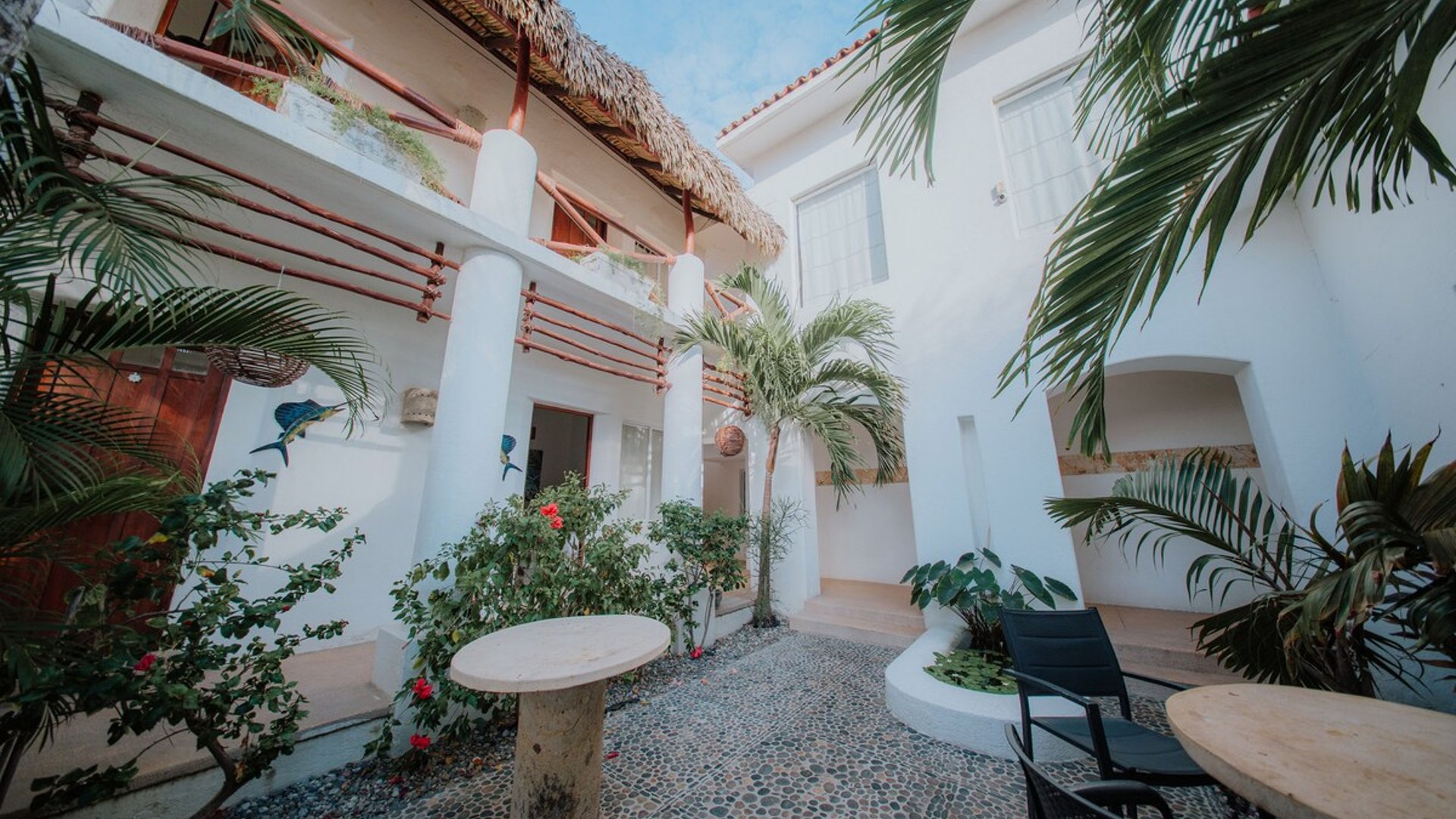 Patio interior empedrado con palmera central, vegetación tropical y fachada blanca de dos niveles con palapa
