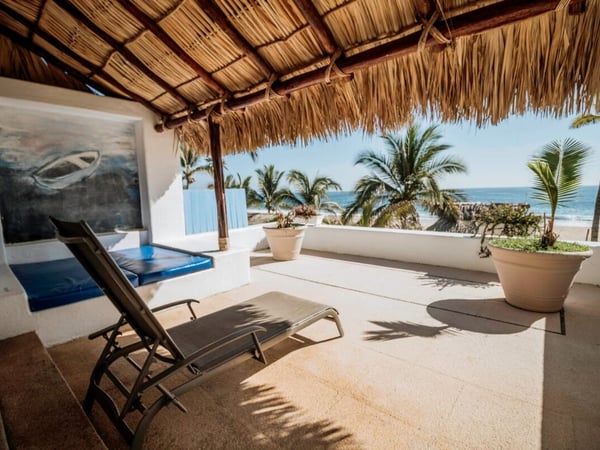 Terraza con palapa y camastro frente al mar, palmeras y playa visibles bajo cielo despejado