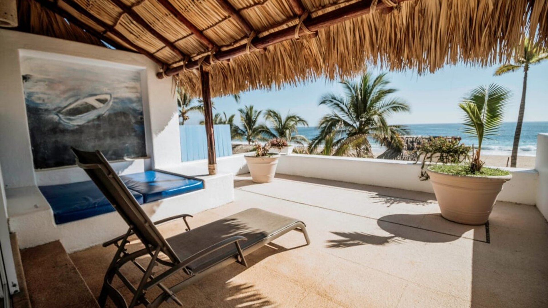 Terraza con palapa y camastro frente al mar, palmeras y playa visibles bajo cielo despejado