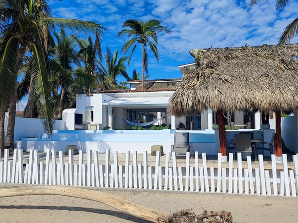 Fachada de villa frente al mar con palapa, hamaca y barda blanca de madera vista desde la playa