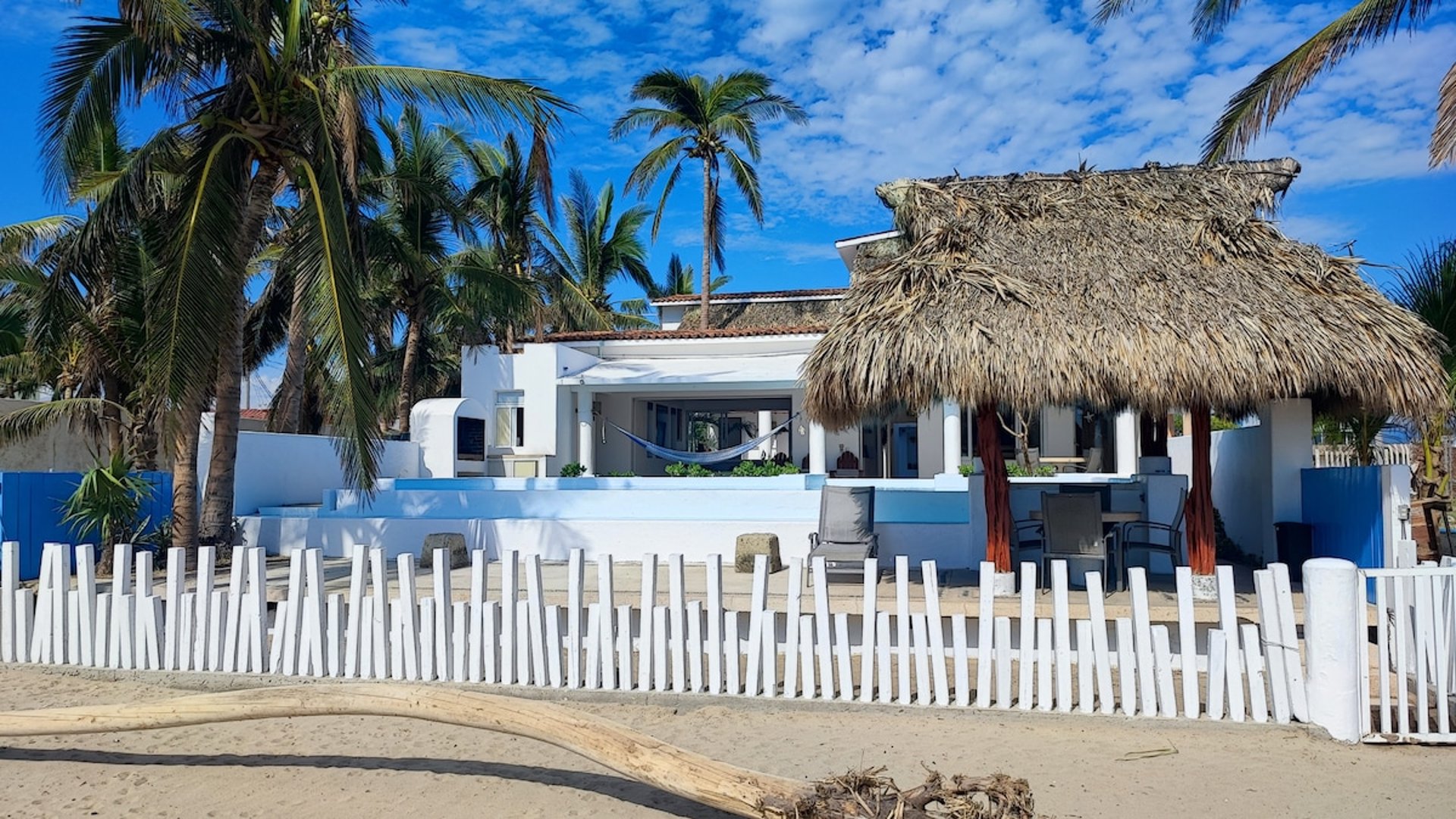 Fachada de villa frente al mar con palapa, hamaca y barda blanca de madera vista desde la playa