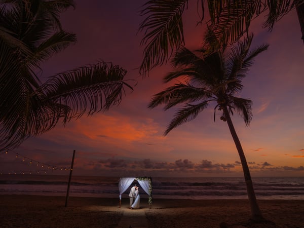 Ceremonia de boda en playa al atardecer con arco decorado bajo palmeras y luces colgantes