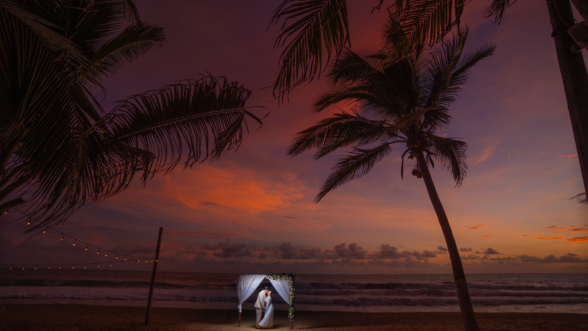 Ceremonia de boda en playa al atardecer con arco decorado bajo palmeras y luces colgantes