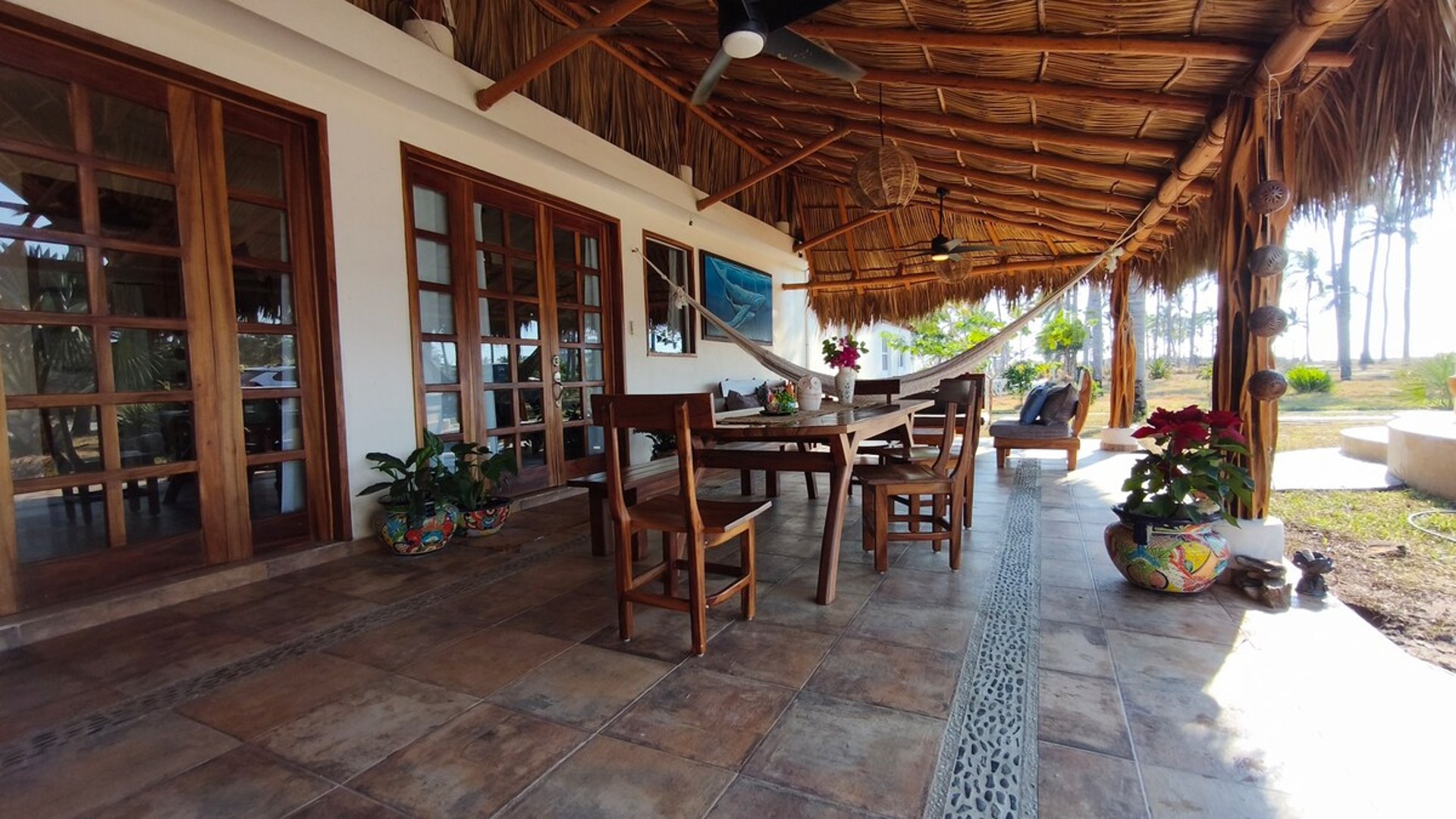 Terraza con palapa, mesa de madera, hamaca y macetas talavera frente a jardín con palmeras al fondo