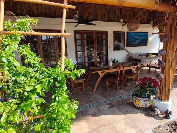 Terraza bajo palapa con comedor de madera, hamaca, vegetación trepadora y decoración artesanal mexicana