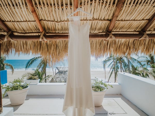 Vestido de novia colgado bajo palapa frente a la playa con macetas decorativas y palmeras