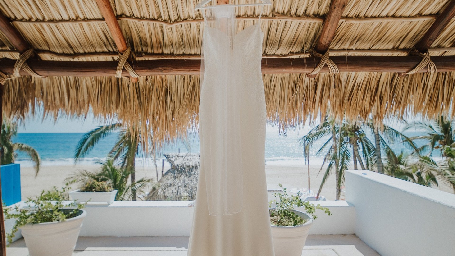 Vestido de novia colgado bajo palapa frente a la playa con macetas decorativas y palmeras
