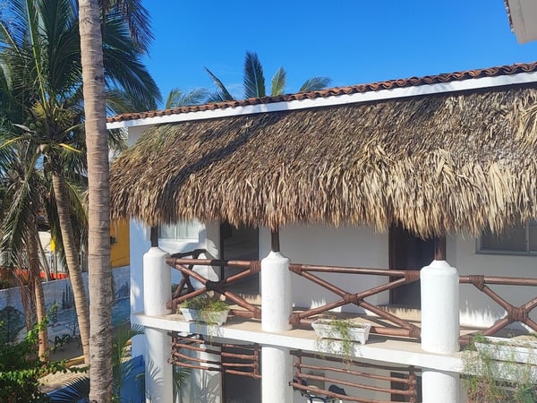 Fachada de cabaña con techo de palapa, barandal de bambú y palmeras bajo cielo azul despejado