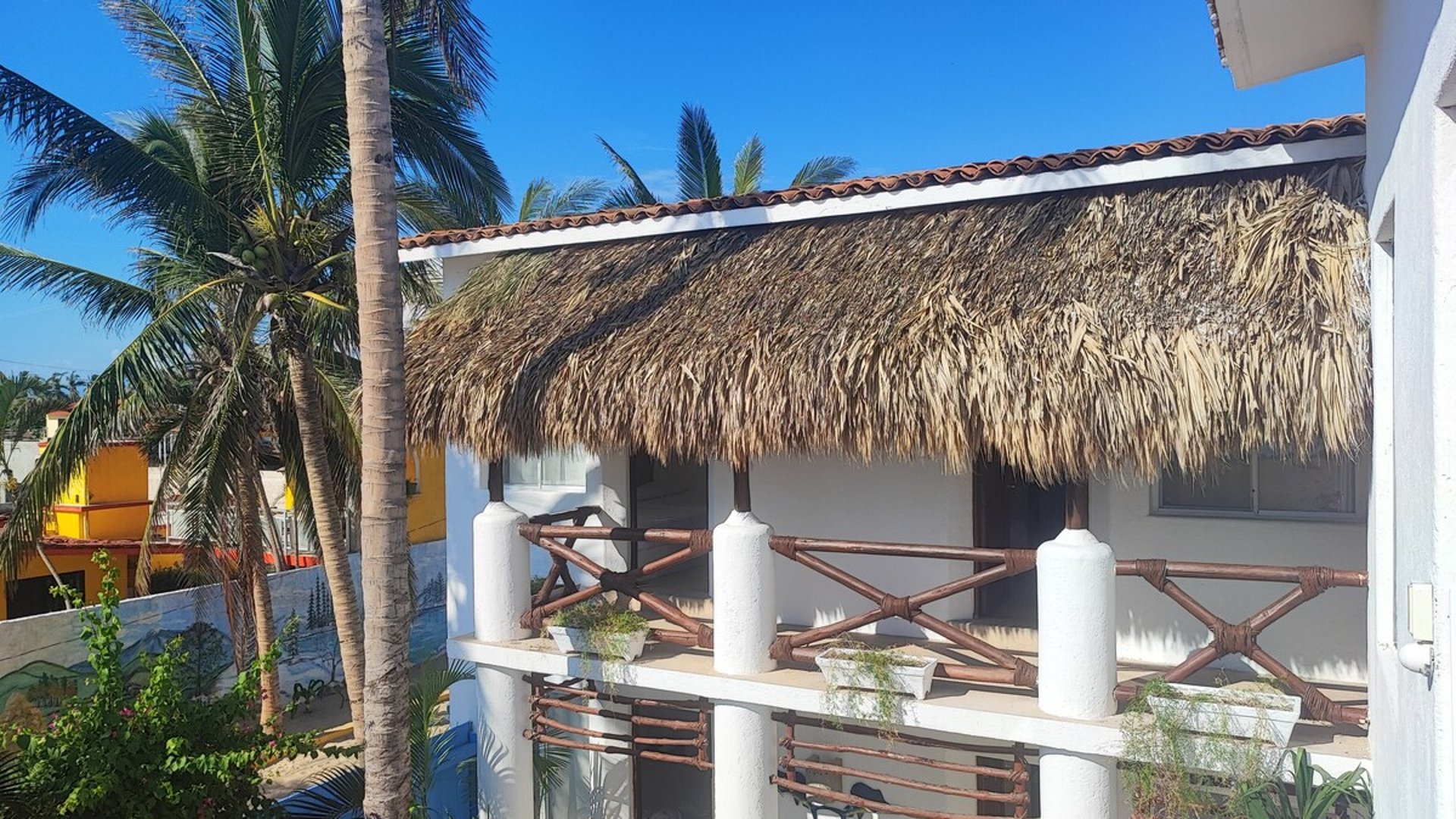 Fachada de cabaña con techo de palapa, barandal de bambú y palmeras bajo cielo azul despejado