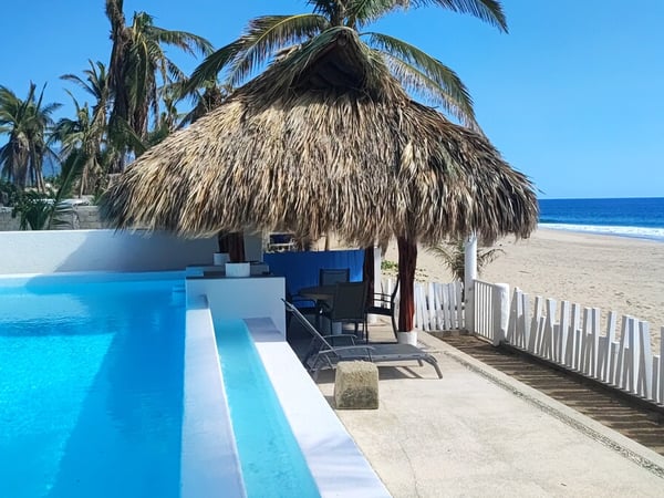 Alberca desbordante junto a palapa con camastros, playa de arena y mar azul al fondo bajo cielo despejado