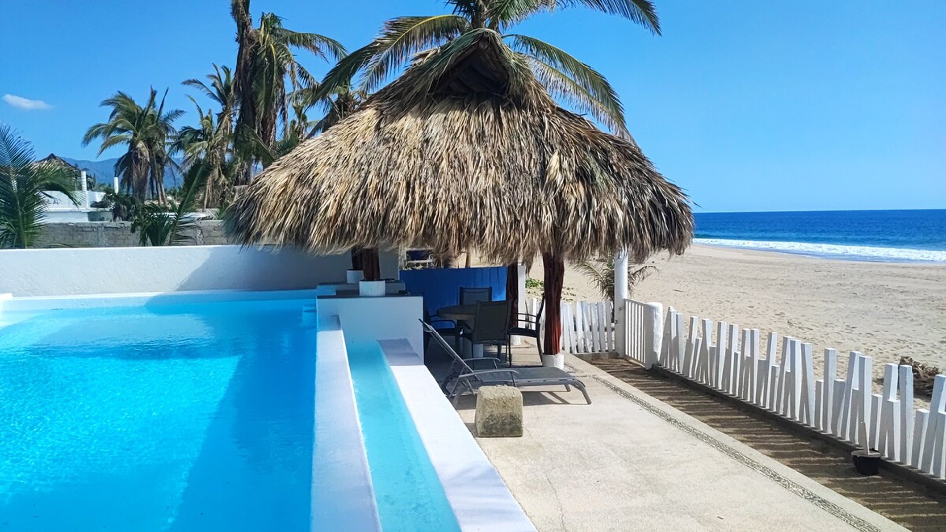Alberca desbordante junto a palapa con camastros, playa de arena y mar azul al fondo bajo cielo despejado