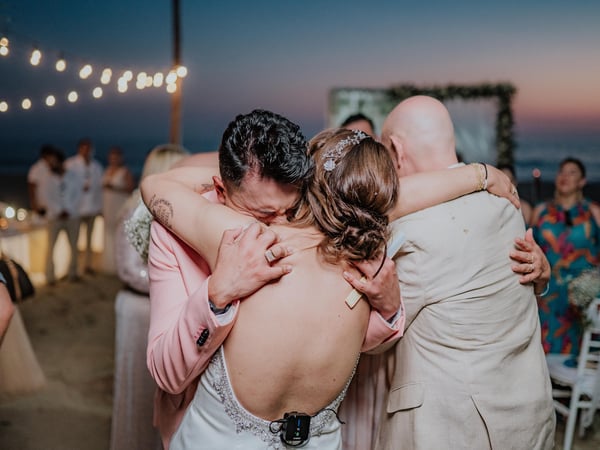 Abrazo emotivo durante recepción de boda en terraza con vista al mar al atardecer y luces colgantes