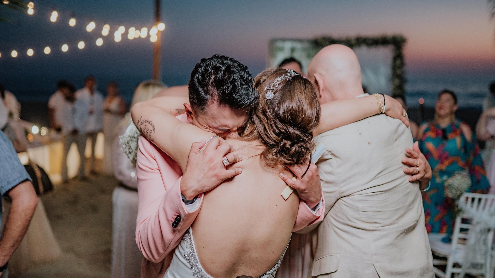 Abrazo emotivo durante recepción de boda en terraza con vista al mar al atardecer y luces colgantes