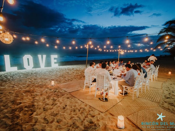 Cena romántica en la playa al anochecer con letras LOVE iluminadas, guirnaldas de luces y grupo de invitados sentados a la mesa