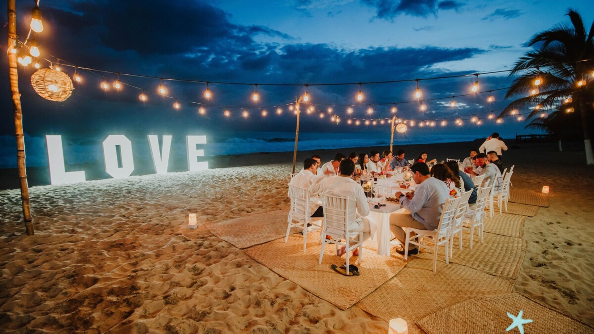 Cena romántica en la playa al anochecer con letras LOVE iluminadas, guirnaldas de luces y grupo de invitados sentados a la mesa