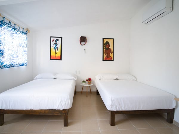 Recámara con dos camas individuales de madera, ropa de cama blanca, arte colorido en paredes y aire acondicionado
