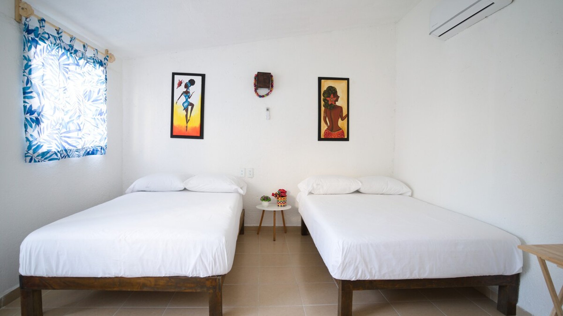 Recámara con dos camas individuales de madera, ropa de cama blanca, arte colorido en paredes y aire acondicionado