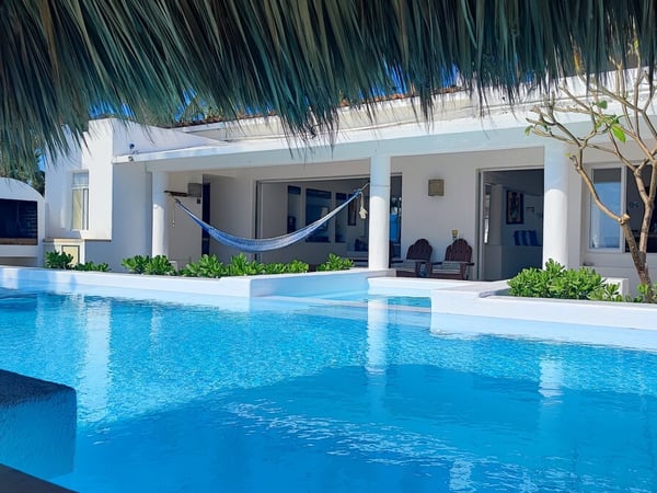 Alberca de aguas cristalinas frente a villa blanca con hamaca y palapa, ambiente tropical luminoso