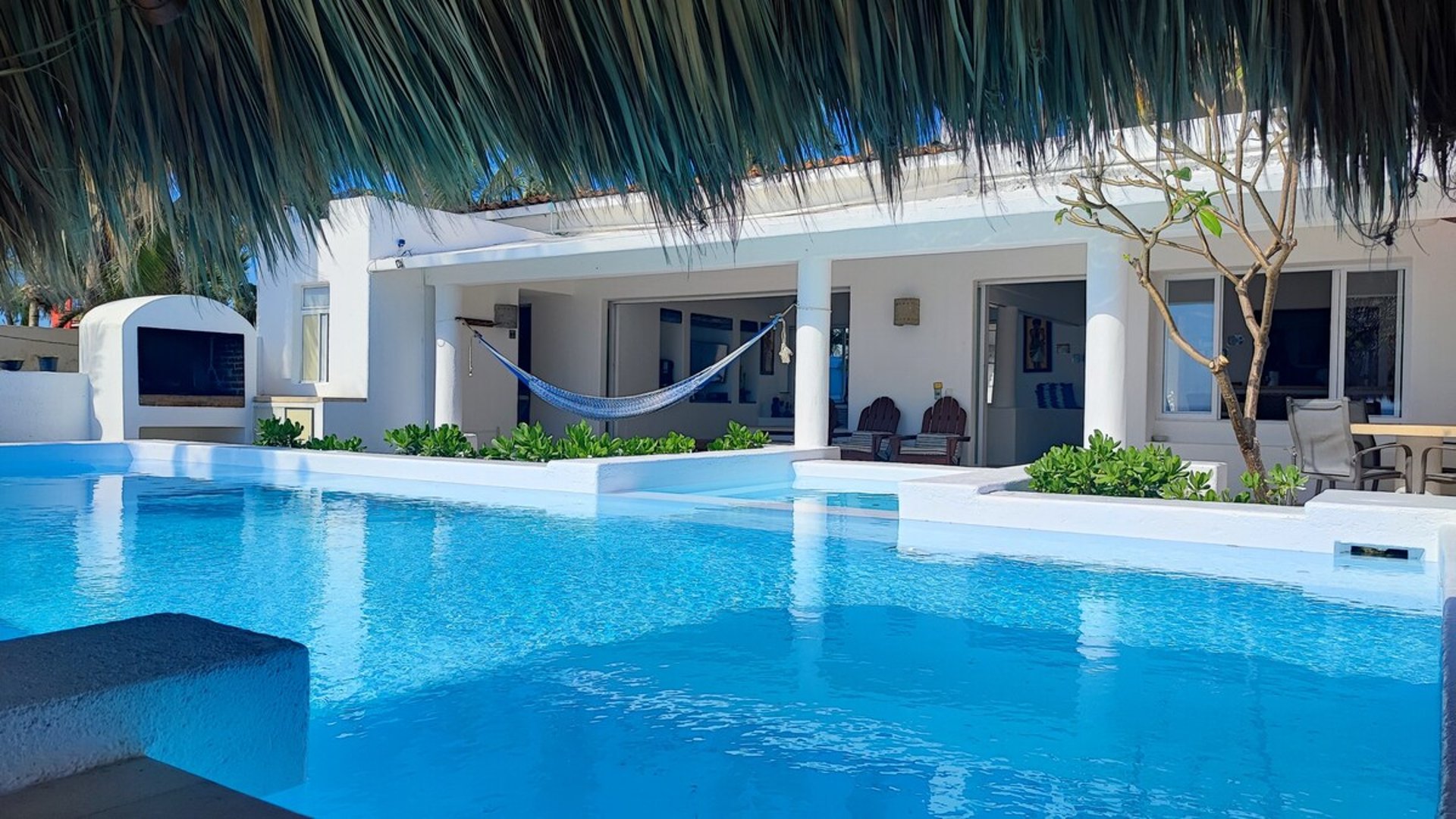 Alberca de aguas cristalinas frente a villa blanca con hamaca y palapa, ambiente tropical luminoso