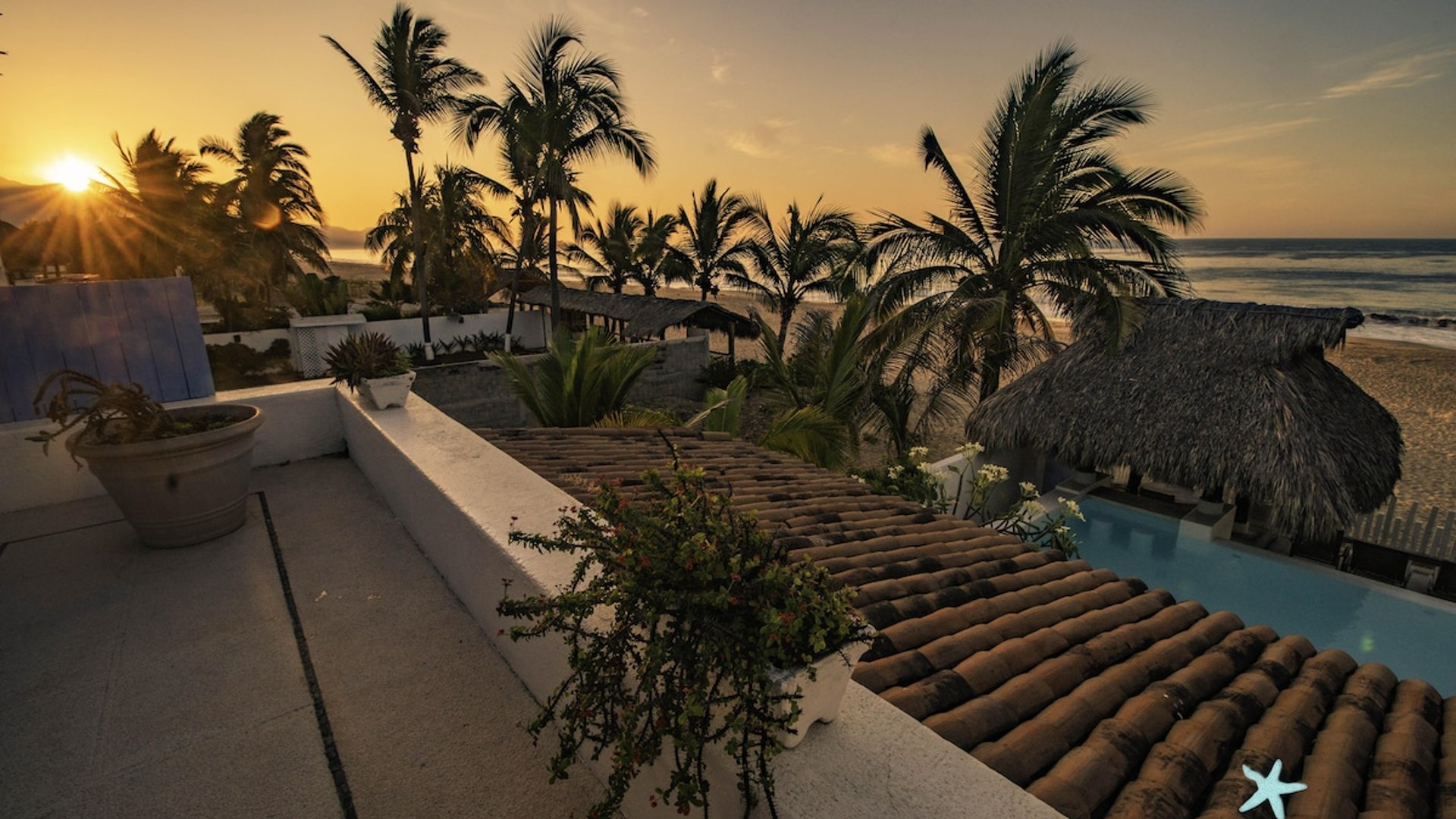 Vista desde terraza al atardecer con palapas, alberca, palmeras y oleaje del Pacífico al fondo