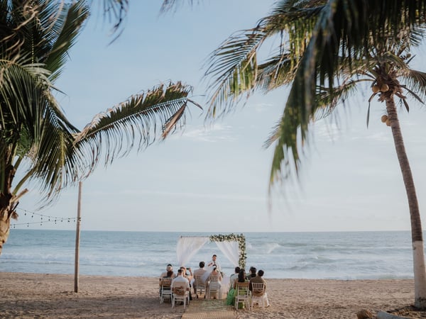 Ceremonia íntima en playa con arco floral blanco frente al mar entre palmeras y luces colgantes