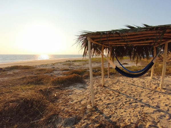 Hamaca azul bajo palapa rústica frente a playa de arena con mar y atardecer dorado al fondo