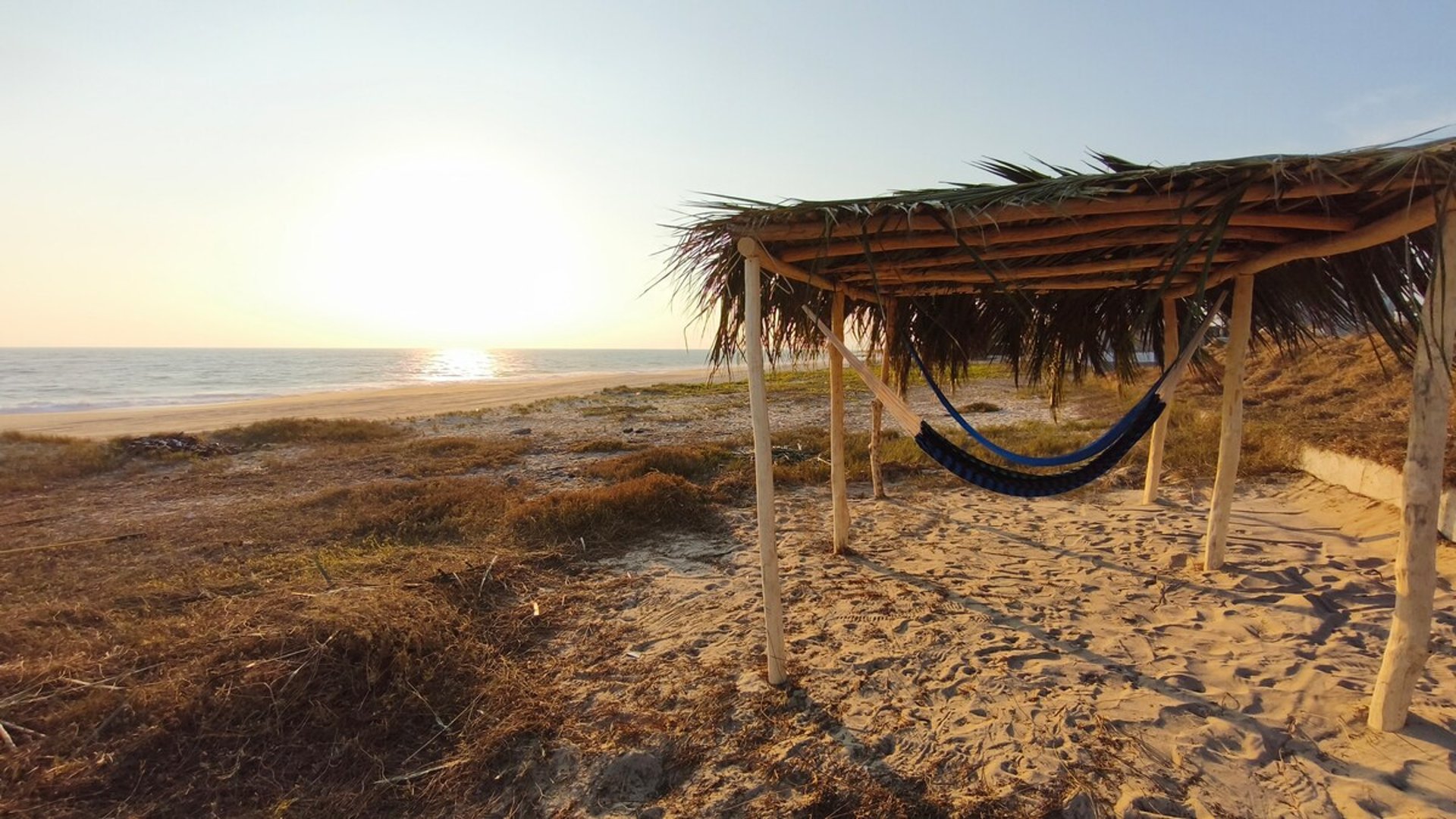 Hamaca azul bajo palapa rústica frente a playa de arena con mar y atardecer dorado al fondo