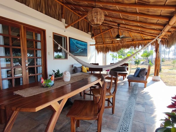 Terraza bajo palapa con mesa de madera, hamaca colgante, sala exterior y vista al jardín con palmeras