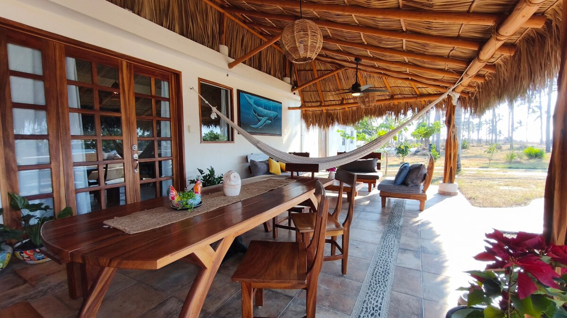 Terraza bajo palapa con mesa de madera, hamaca colgante, sala exterior y vista al jardín con palmeras