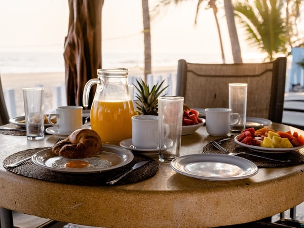 Mesa redonda de piedra con desayuno frente a la playa: jugo, fruta, croissant y huevos con palmeras al fondo